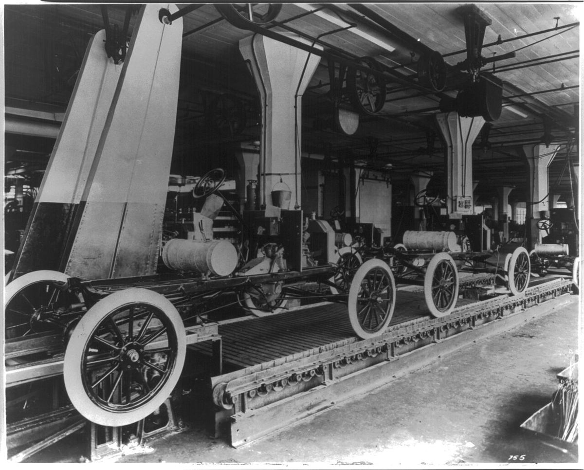 A black-and-white photo of early automobile chassis with spoked wheels moving down a factory assembly line