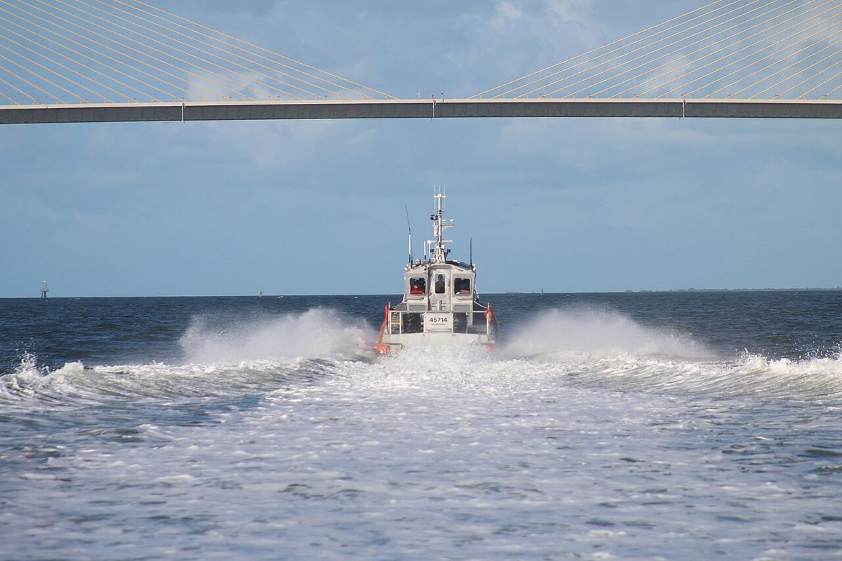 A Coast Guard response boat speeds across blue Florida water, throwing a white wake beneath a long bridge