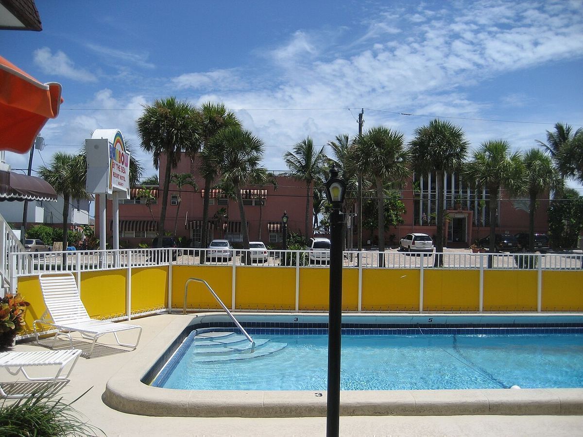 An empty outdoor swimming pool with lounge chairs, a yellow wall and palm trees under a blue Florida sky