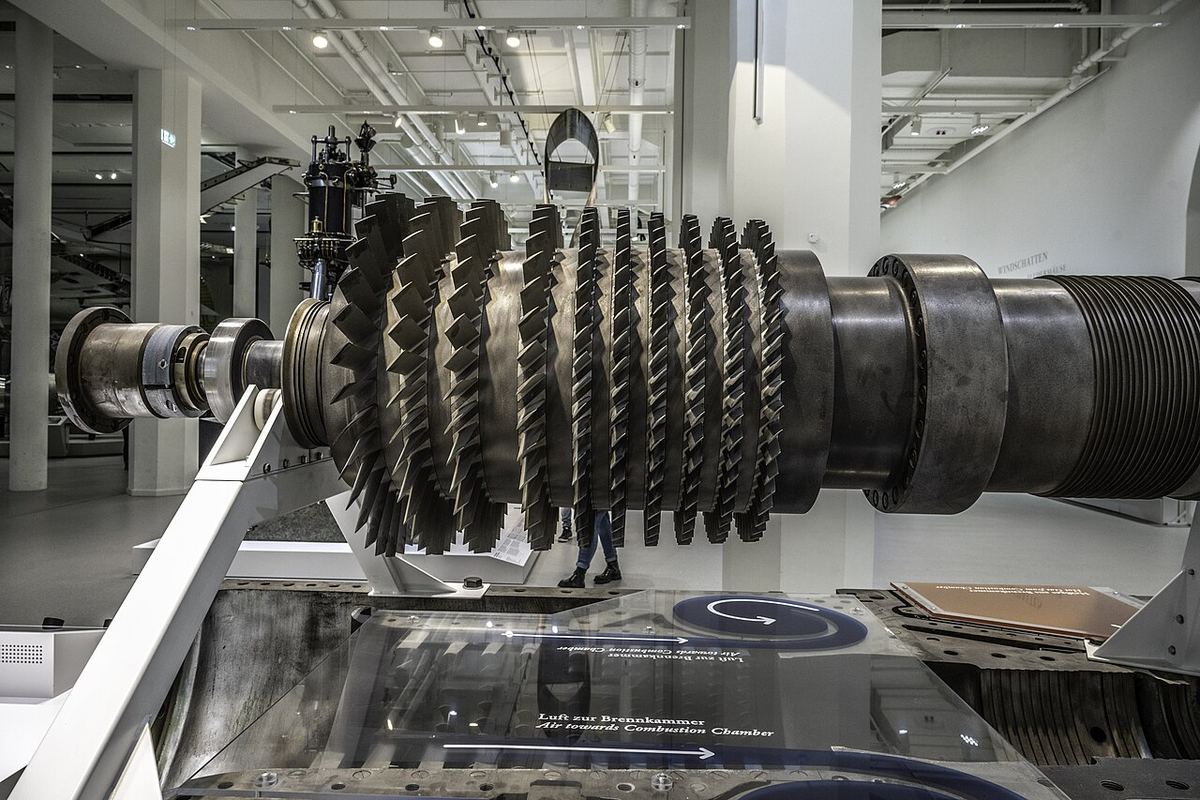 The bladed steel compressor rotor of a large gas turbine rests on a stand in a bright museum hall