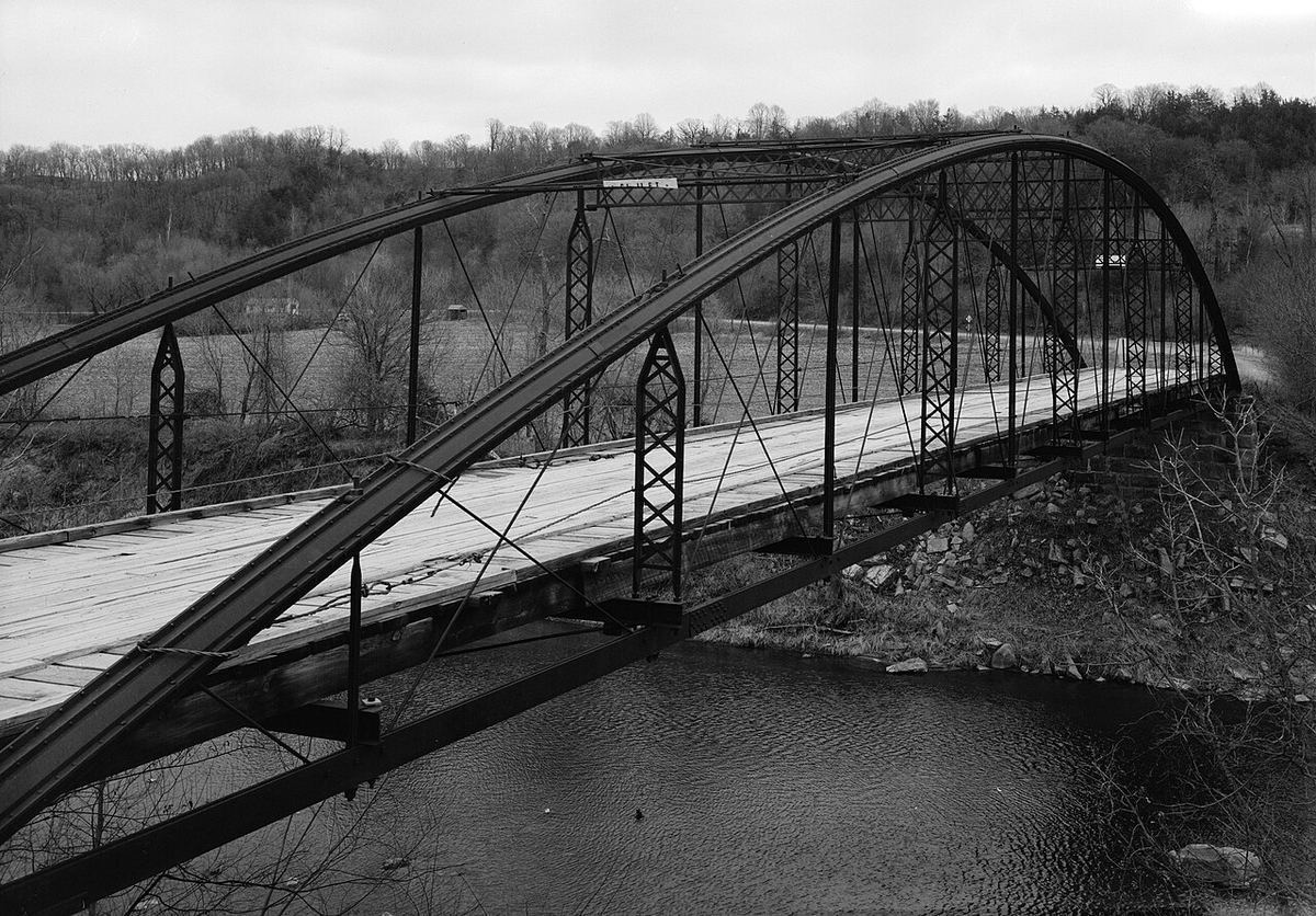 A black-and-white view of the Kern Bridge's iron bowstring truss arching over the Blue Earth River among bare trees