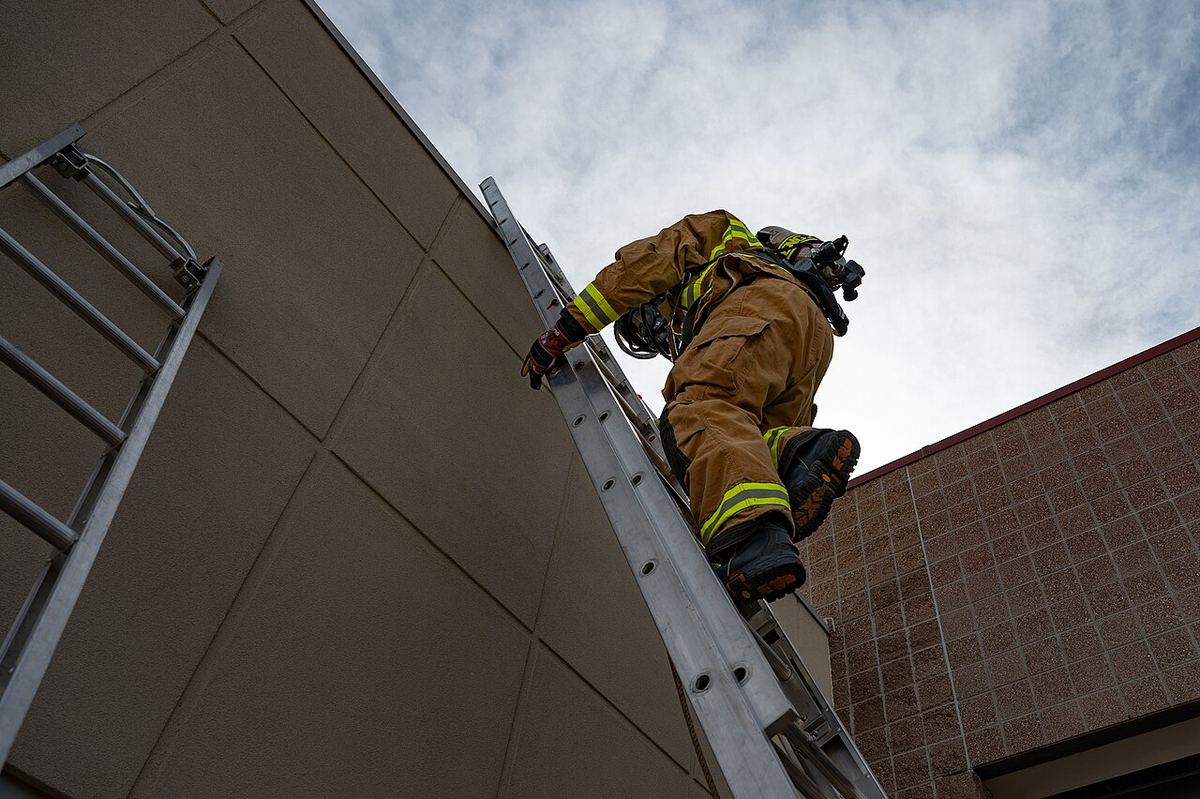 Seen from below, a firefighter in full turnout gear climbs an aluminum ladder against a brick building under a cloudy sky