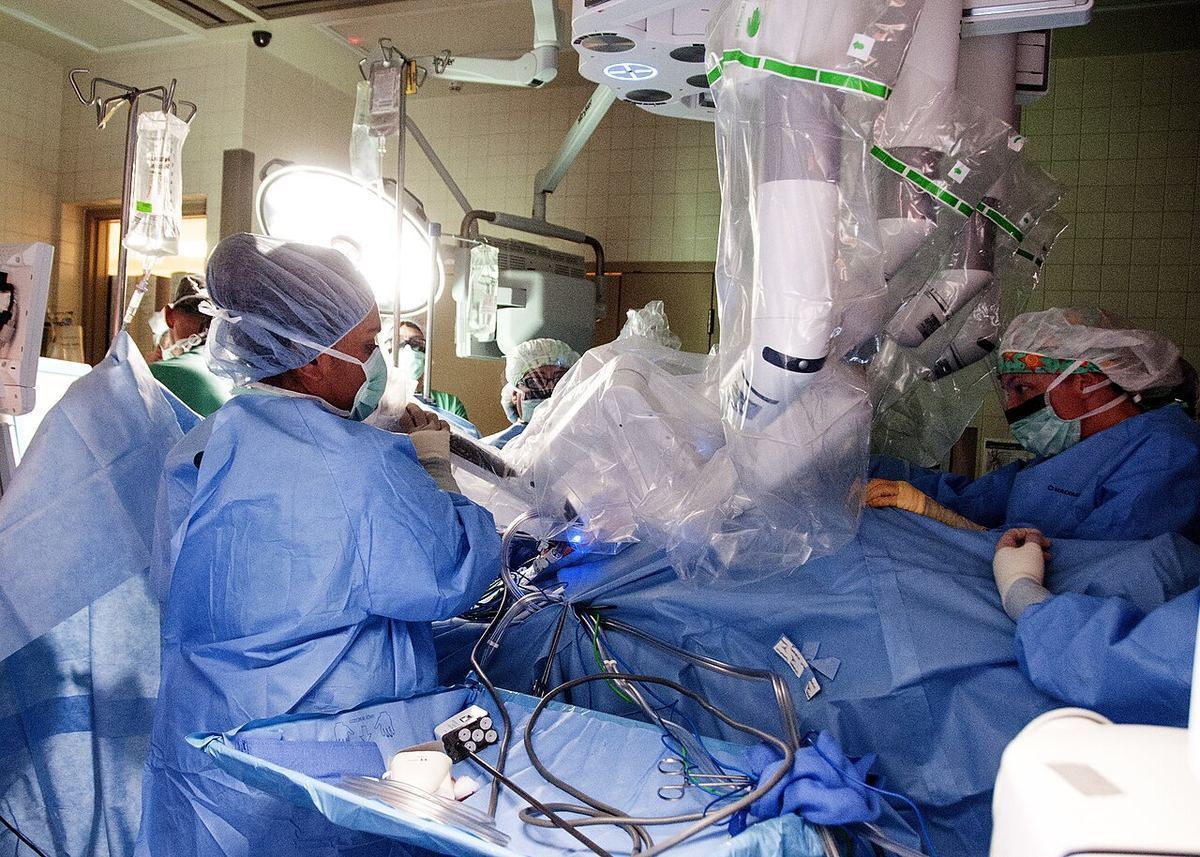 Masked surgeons in blue gowns work beside the white arms of a robotic surgical system in a bright operating room