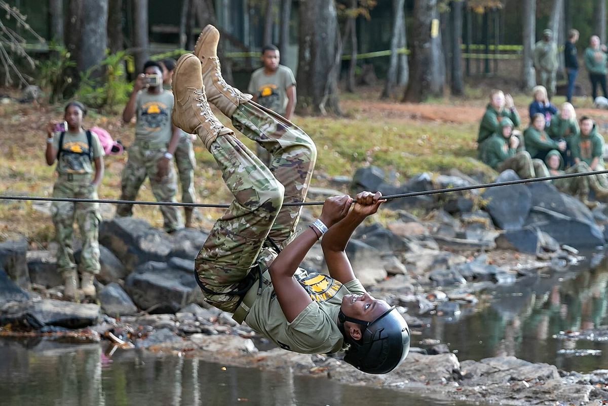 A JROTC cadet in camouflage hangs upside down from a single rope stretched over a rocky creek while other cadets watch from the bank