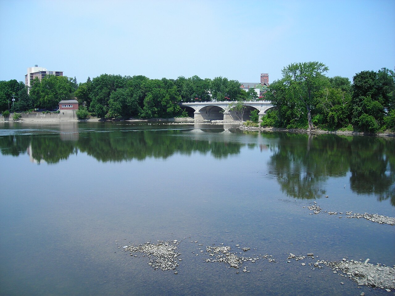 The Susquehanna River on a calm, sunny summer day in Binghamton, N.Y., lined with green trees and an arched stone bridge