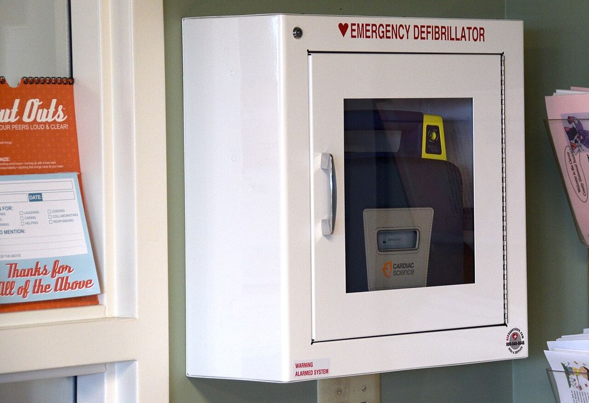 A white wall cabinet labeled Emergency Defibrillator holding an automated external defibrillator