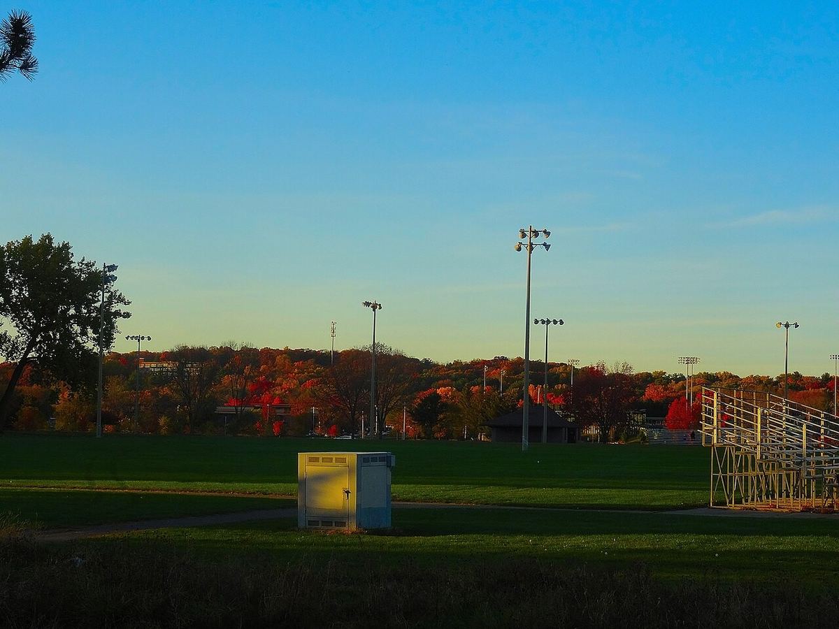 Light towers and bleachers stand over a quiet ballfield at sunset, with red and orange autumn trees behind