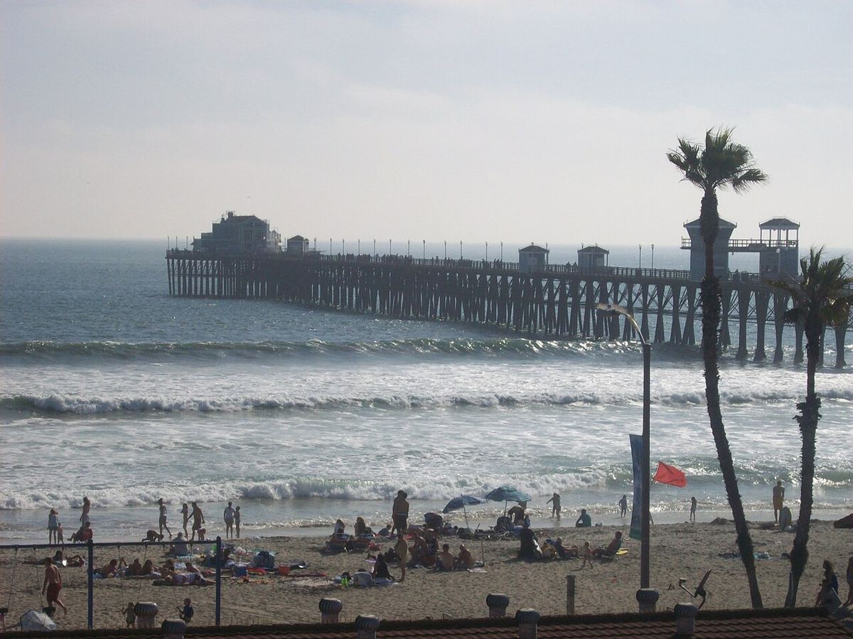 Waves rolling toward a sandy beach beside the long wooden Oceanside Pier, with palm trees and distant beachgoers