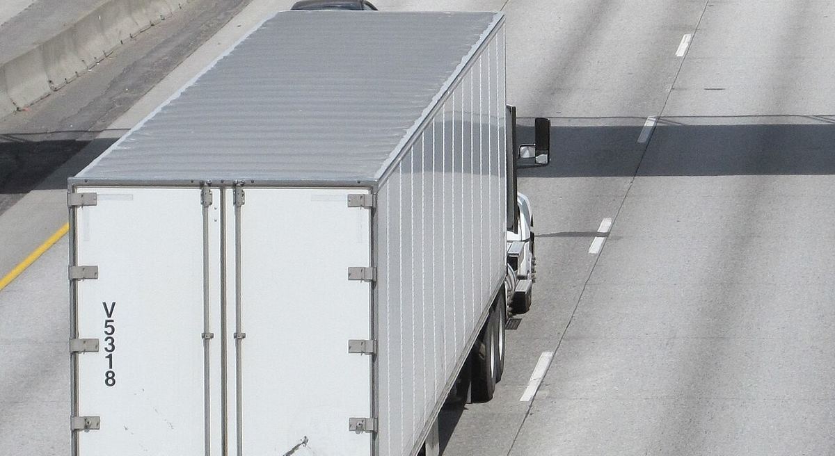 A white 53-foot dry van semi-trailer rolls down a concrete freeway, seen from above