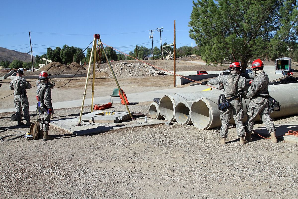 Rescuers in red helmets set up a tall metal tripod and rope lines beside a row of large concrete pipes during a training drill