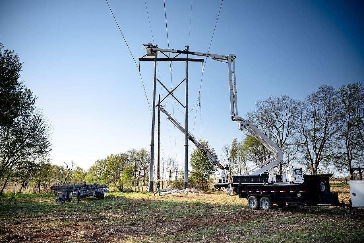 Bucket trucks raising a new power line across a green rural field under a clear spring sky