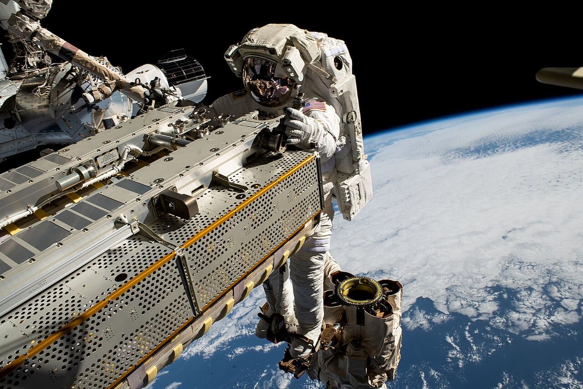 A spacesuited astronaut works beside a space station truss segment high above clouds and the blue curve of Earth
