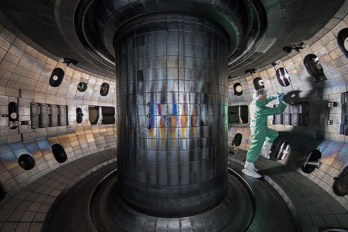 Inside the DIII-D fusion reactor's doughnut-shaped chamber, with tiled walls around a central column and a technician in a white protective suit